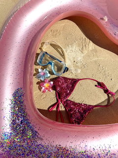 Pink pool float on sand with snorkel mask, flower hair clip, glitter, and pink sequined bikini