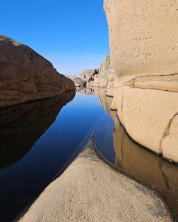 Smooth granite boulders surrounding a still tidal pool with perfect reflections under blue sky