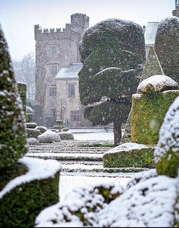 Snow-covered formal garden with topiary leading to a stone castle with tower in winter snowfall