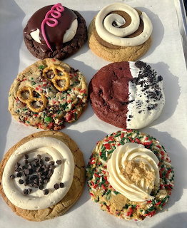 Six large decorated cookies with various toppings including frosting, sprinkles, chocolate chips, and pretzels on parchment paper.