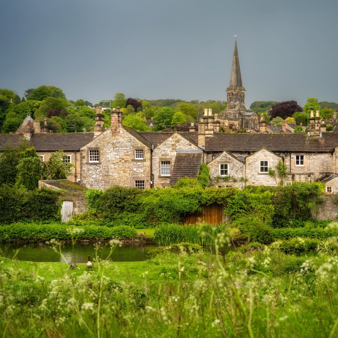 Stone village houses with a church spire behind green fields and trees