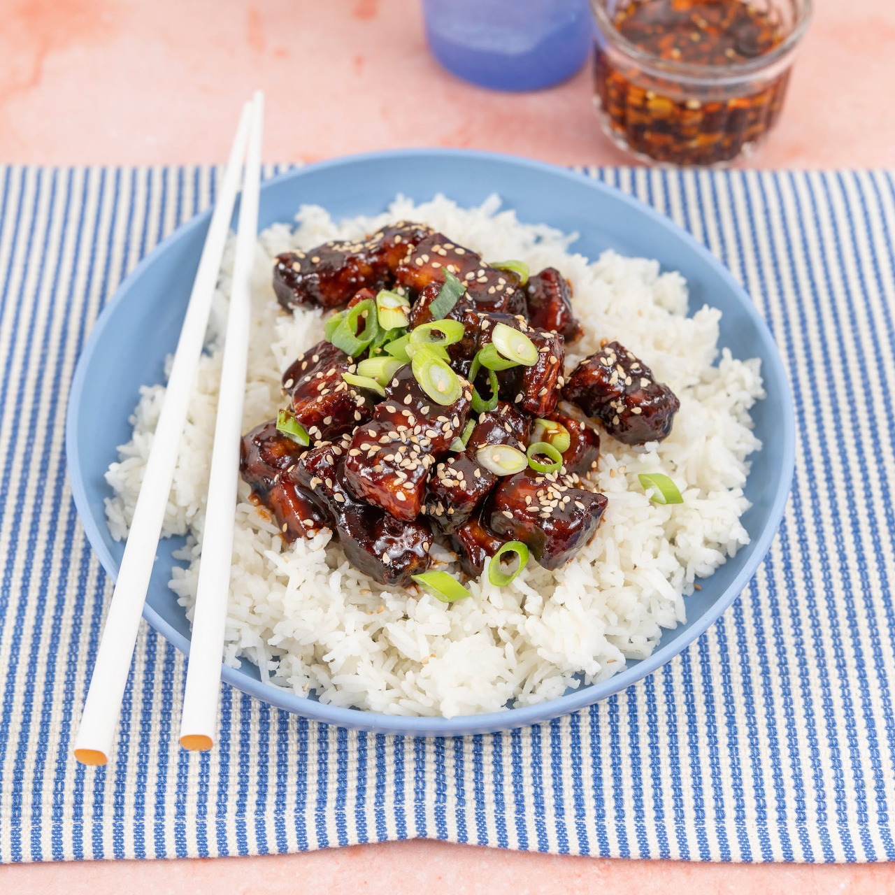 Blue bowl of white rice topped with glazed beef, sesame seeds, and green onions beside chopsticks