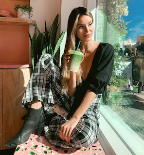 Woman in plaid outfit holding green smoothie by sunny window with plants
