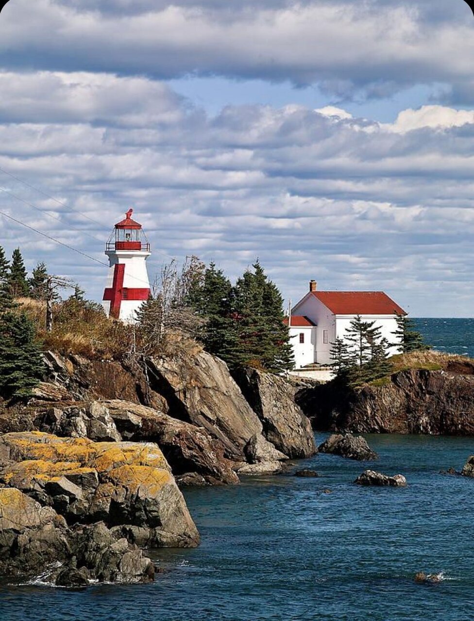 Red and white lighthouse on rocky coastal cliff with evergreen trees and blue ocean water