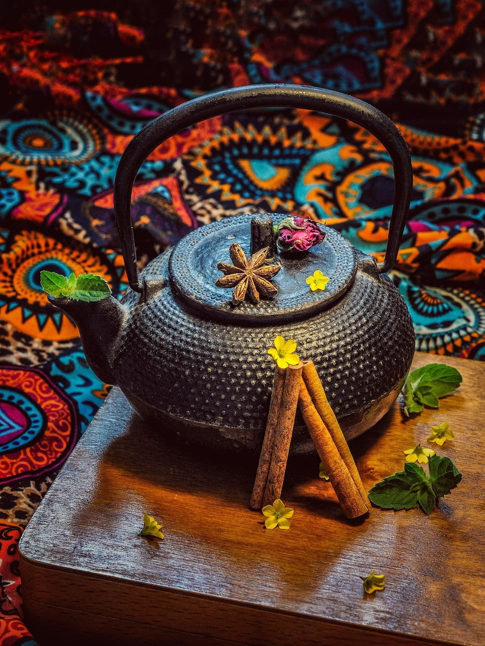 Black cast iron teapot on wooden serving tray with cinnamon sticks and mint against vibrant patterned fabric