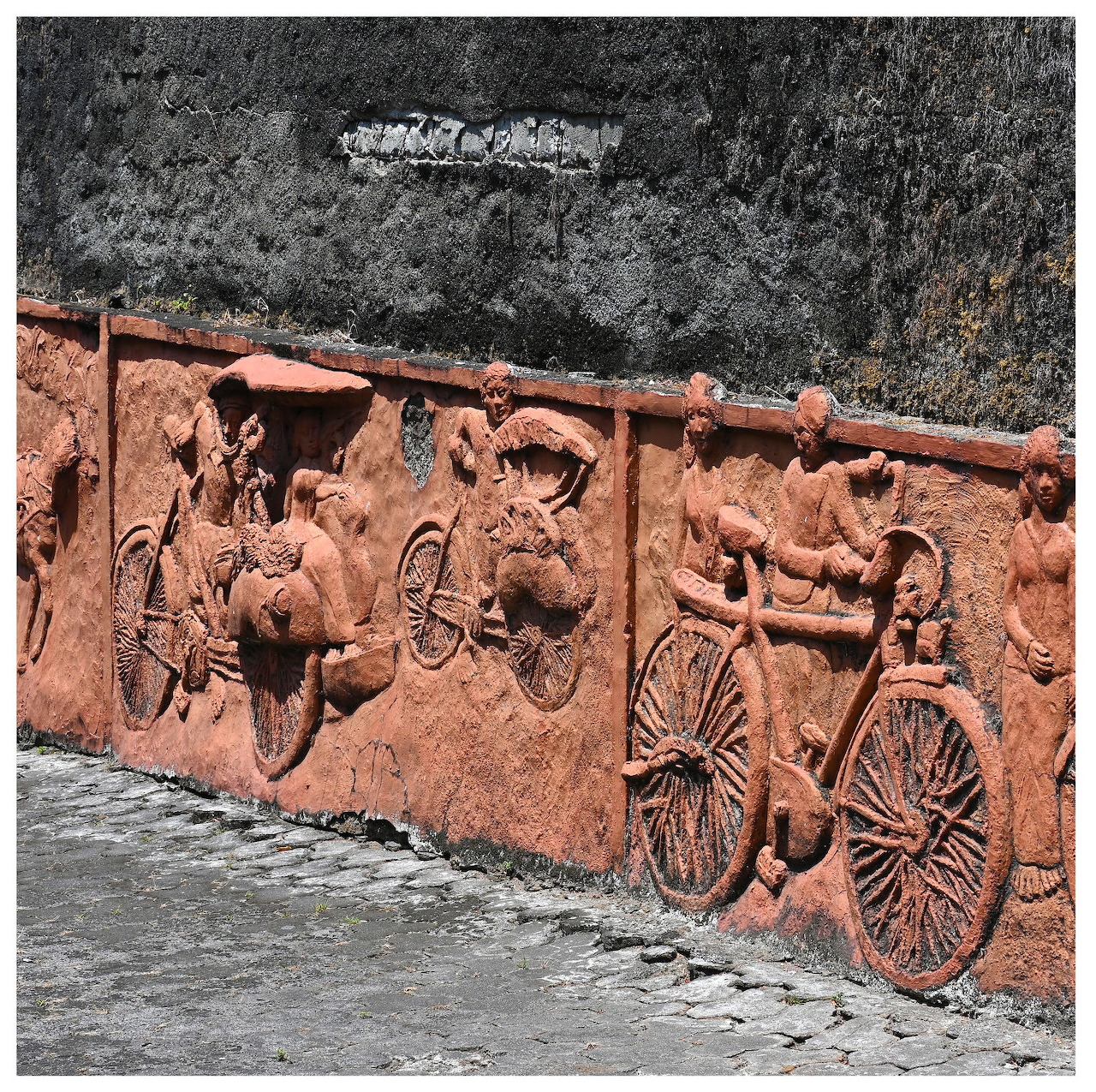 Terracotta wall relief of people riding bicycles against a dark textured stone wall