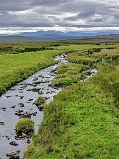 Winding stream through green moorland with distant mountains and cloudy sky in Sutherland, Scotland