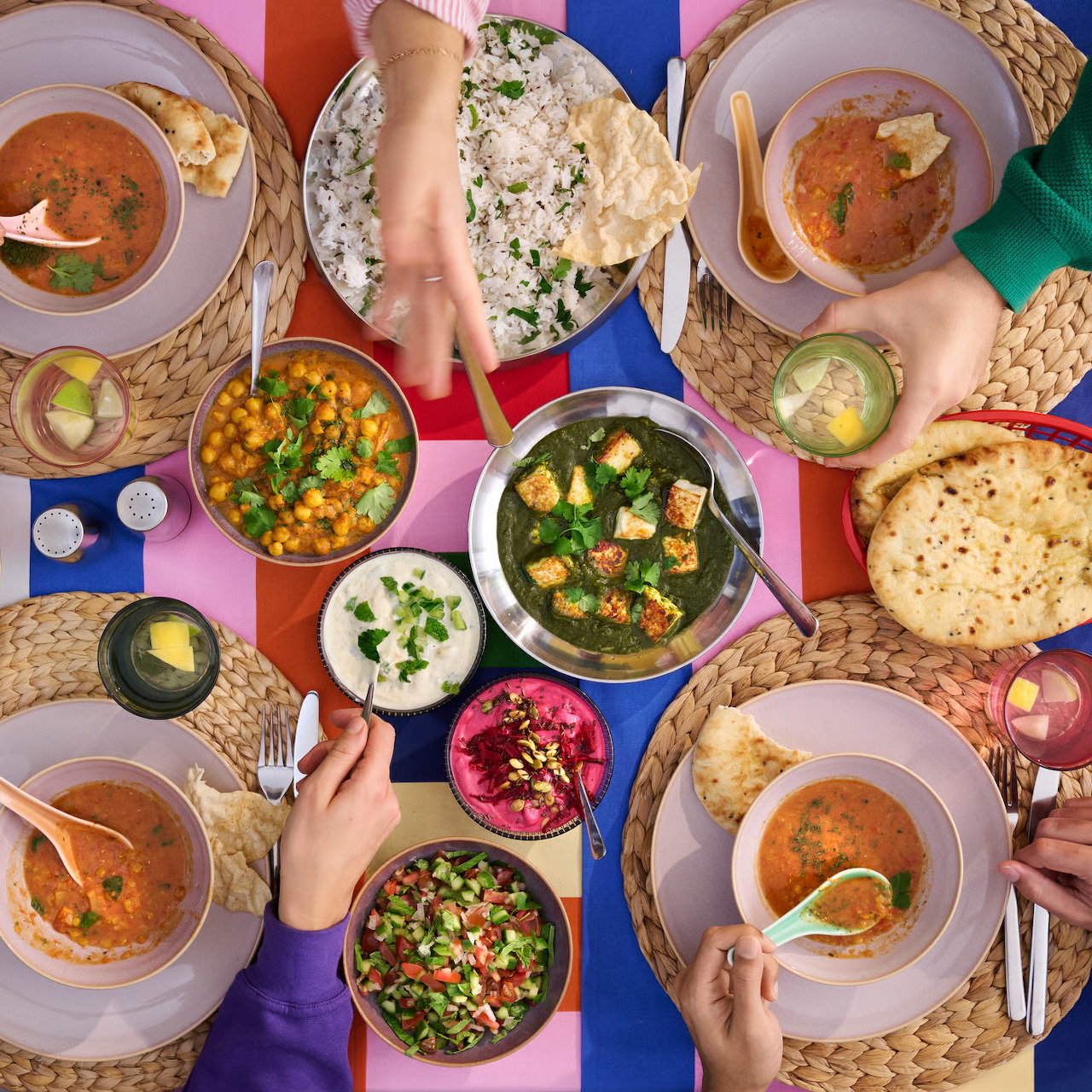 Overhead view of Indian curry dishes, naan bread, and rice arranged on a colorful table with hands visible