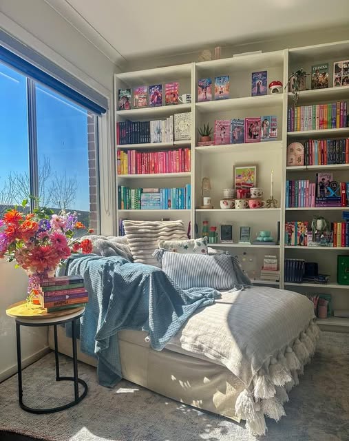 Sunlit reading nook with a daybed, blue blanket, flowers, and bookshelves beside a window