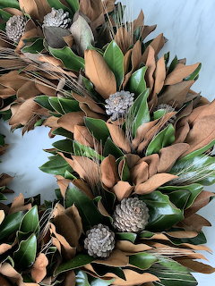 Rustic fall wreath with brown magnolia leaves, green foliage, and silver pine cones on light blue background