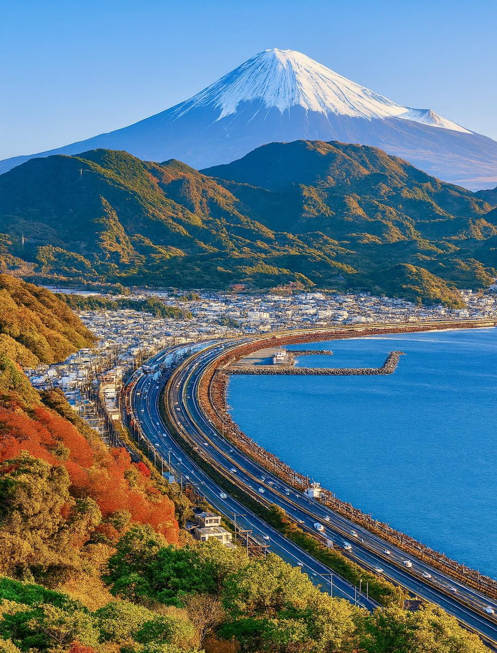 Snow-capped Mt. Fuji towering over a coastal highway, bay, and autumn-colored hills in Japan