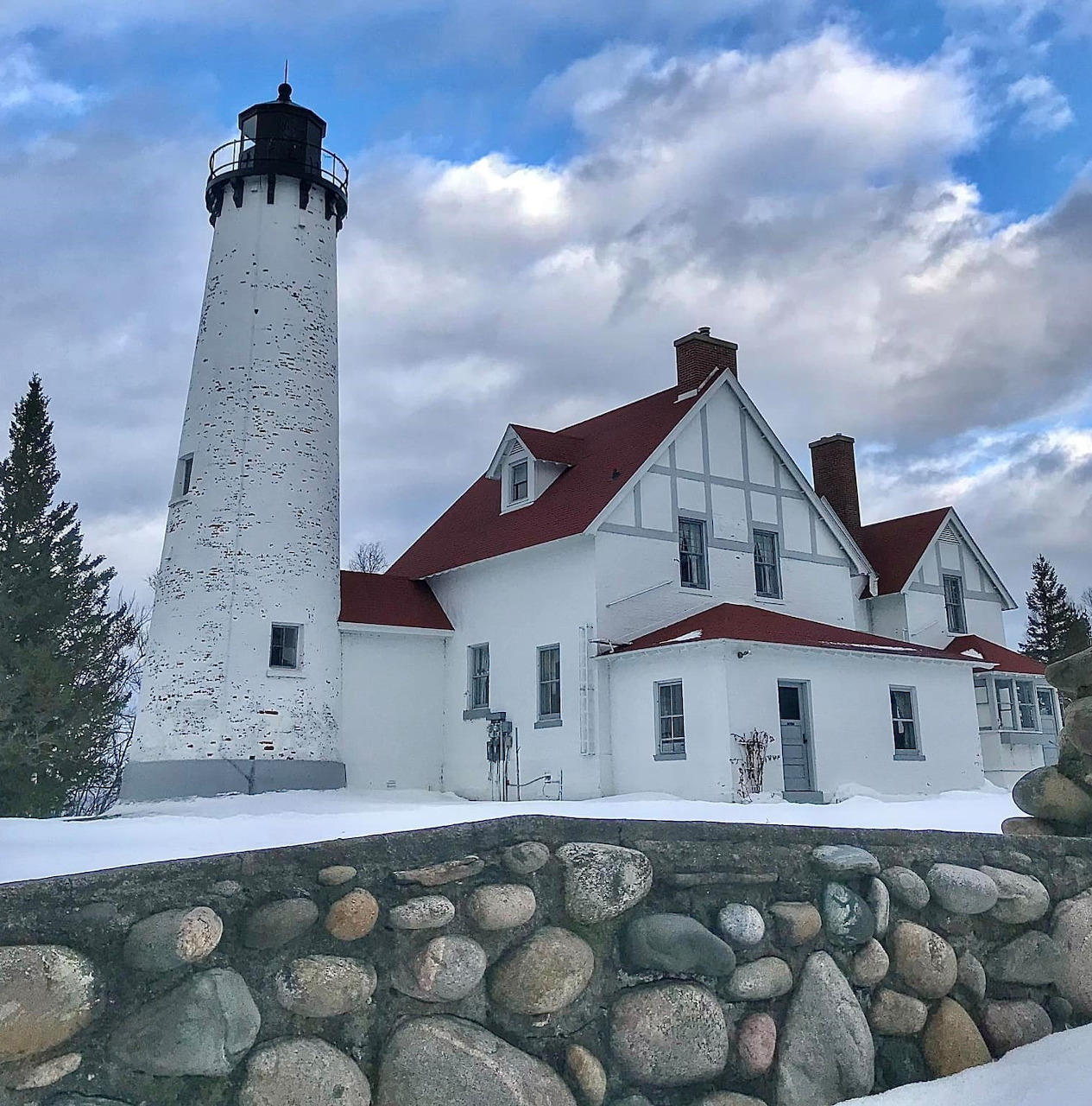 White lighthouse tower and keeper's house with red roof behind stone wall under blue sky with clouds