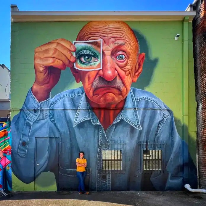 Street art mural of elderly man holding glittery eye mask to face on green wall