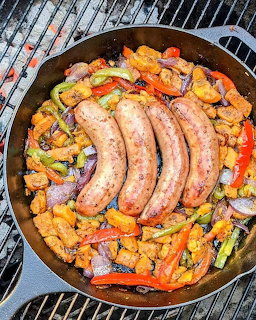 Cast iron skillet on a grill with four sausages surrounded by colorful roasted peppers and onions
