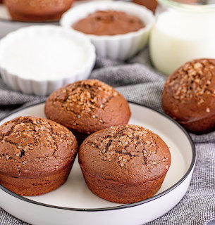 Three chocolate muffins on white plate with milk glass and cocoa powder in background