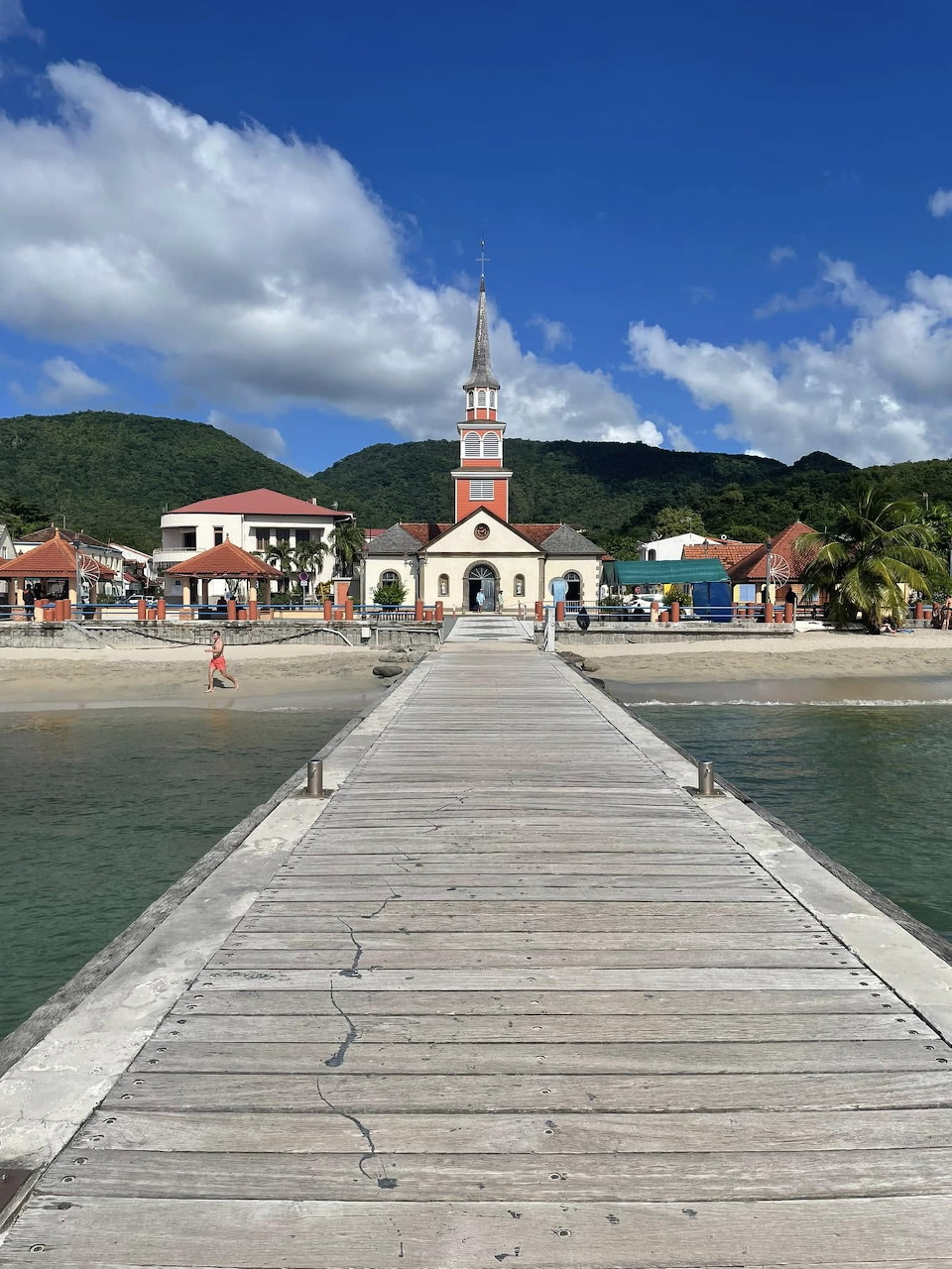 Wooden pier extending toward a red and white church on a Caribbean beach with palm trees and green hills