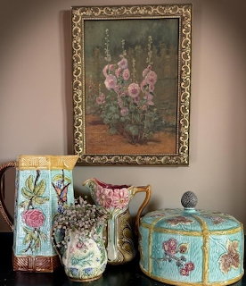 Majolica ceramic pitchers and a lidded jar below a framed hollyhock painting on a wall
