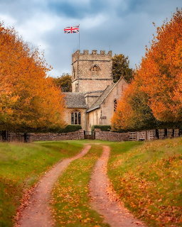 Stone English church with Union Jack flag framed by orange autumn trees along a country lane