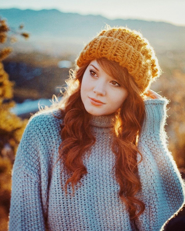 Young redhead woman in yellow beanie and blue sweater outdoors with autumn mountain scenery