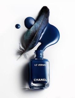 Dark blue nail polish bottle with spilled polish, a feather, and a small droplet on a light background.