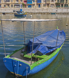Blue covered traditional boat mored in calm green harbor water with Maltese buildings behind