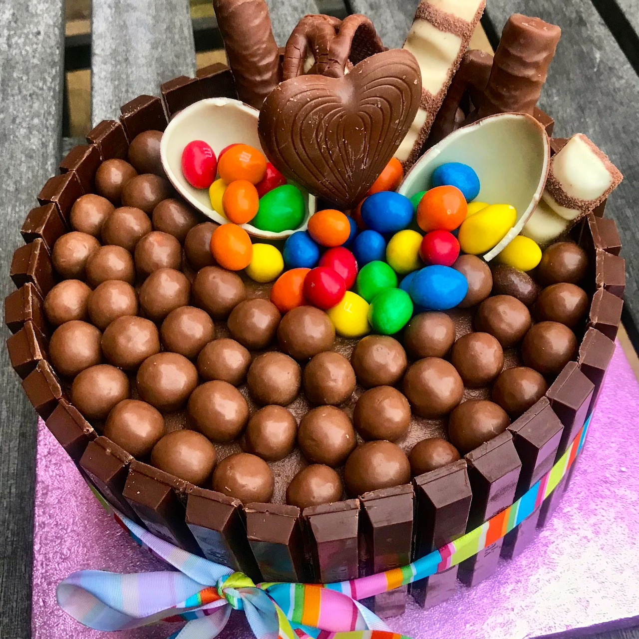 Round chocolate cake topped with chocolate candies, wafer rolls, heart decoration, and colorful candy pieces