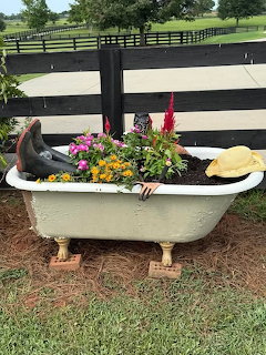 Old clawfoot bathtub flower planter with boots and a straw hat beside a black fence