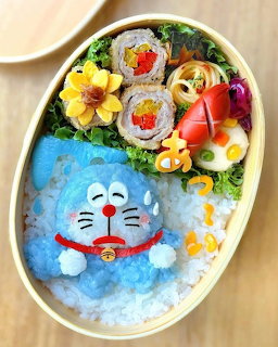 Oval bento box with Doraemon character made from blue rice, sushi rolls, flowers, and colorful food decorations