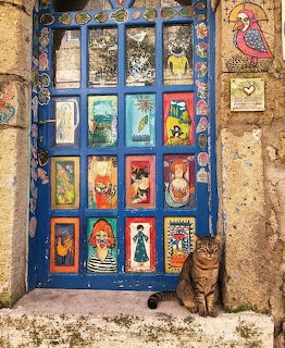 Tabby cat sitting next to blue door decorated with colorful mosaic tile art panels