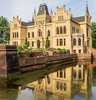 Yellow castle with towers and arched windows reflected in surrounding moat, flower pots on balustrade