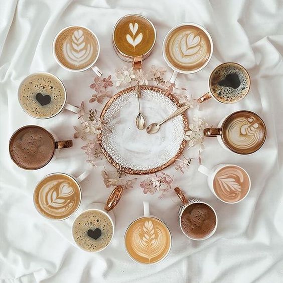 Coffee cups with latte art arranged in clock formation around rose gold timepiece on white fabric