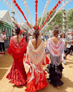 Three women in red, orange, and pink flamenco dresses walking at the Feria de Sevilla with lanterns above