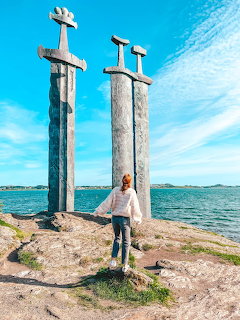 Woman standing in front of three giant sword monuments at Sverd i Fjell, Norway, with ocean and blue sky