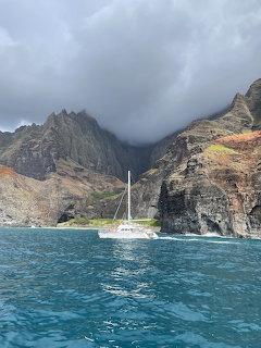 White sailboat on turquoise ocean water with tall dramatic cliffs and cloudy sky in background