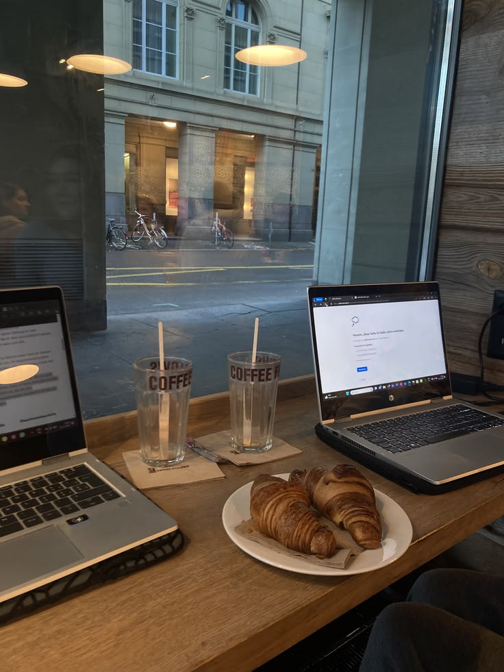 Two open laptops and a plate of croissants on a cafe table by a window with a street view outside