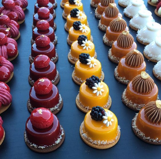 Rows of colorful French mini pastries including red, orange, brown and white desserts on a dark surface