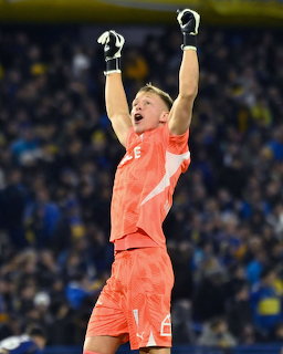 Goalkeeper in orange kit celebrating with arms raised at a football stadium at night