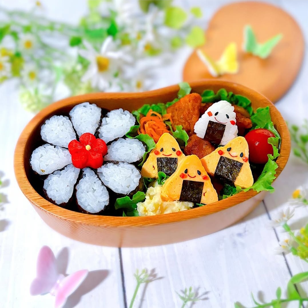 Wooden bento box with flower-shaped rice balls, character onigiri, egg rolls, and lettuce on floral tablecloth