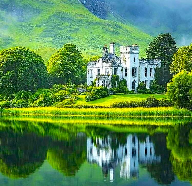 White abbey by a lake with green hills, trees, and a mirrored reflection