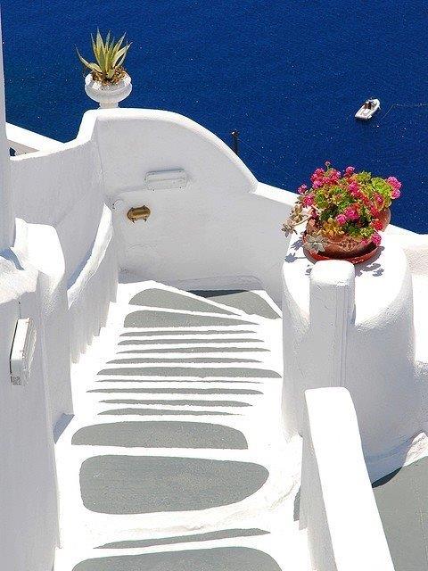 White steps and walls in Santorini above blue sea, with pink flowers and a small boat