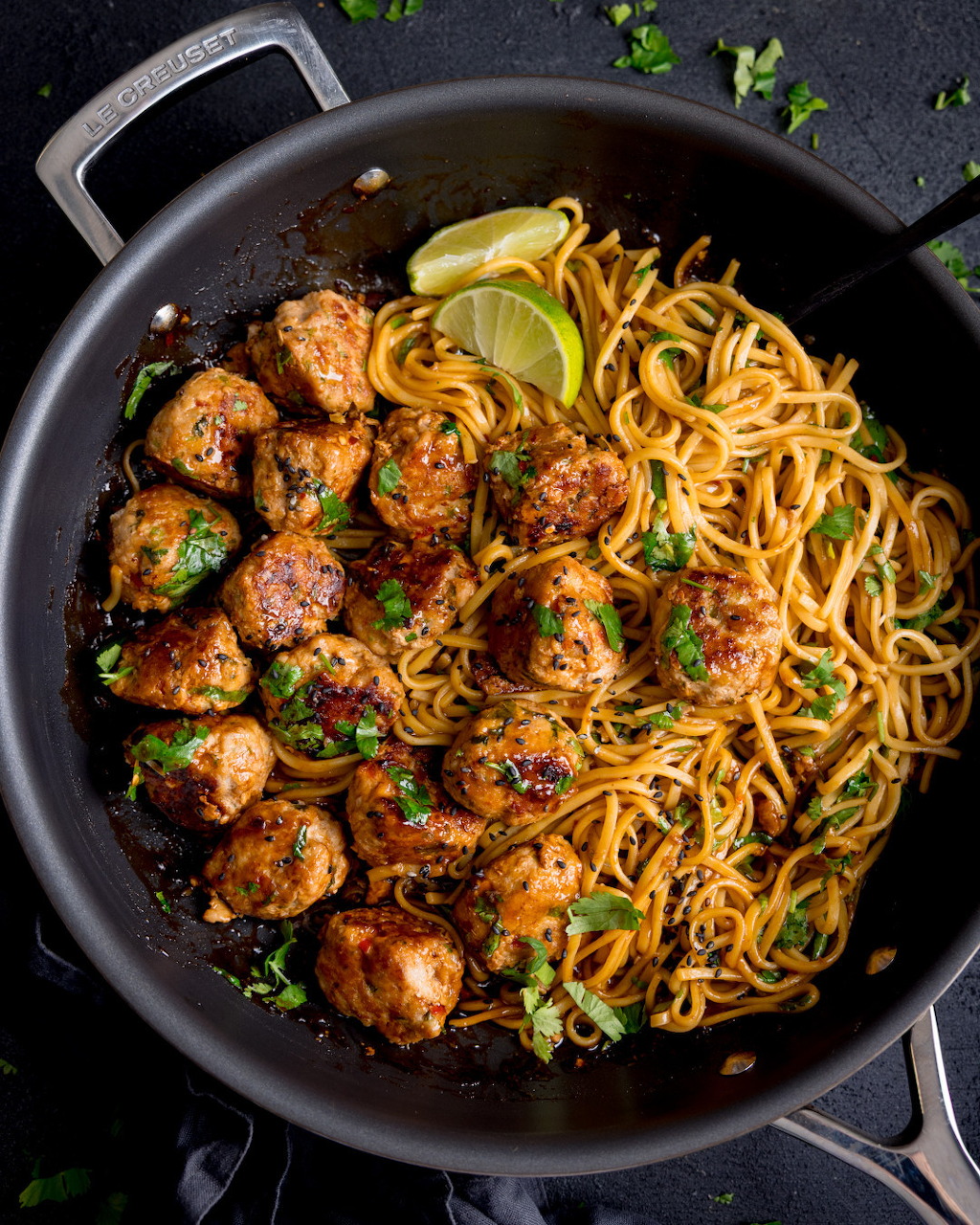 Top-down view of a wok with noodles, chicken meatballs, cilantro, and lime wedges