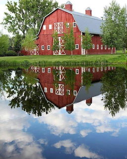 Large red barn with white windows and trim reflected in a still pond surrounded by green trees