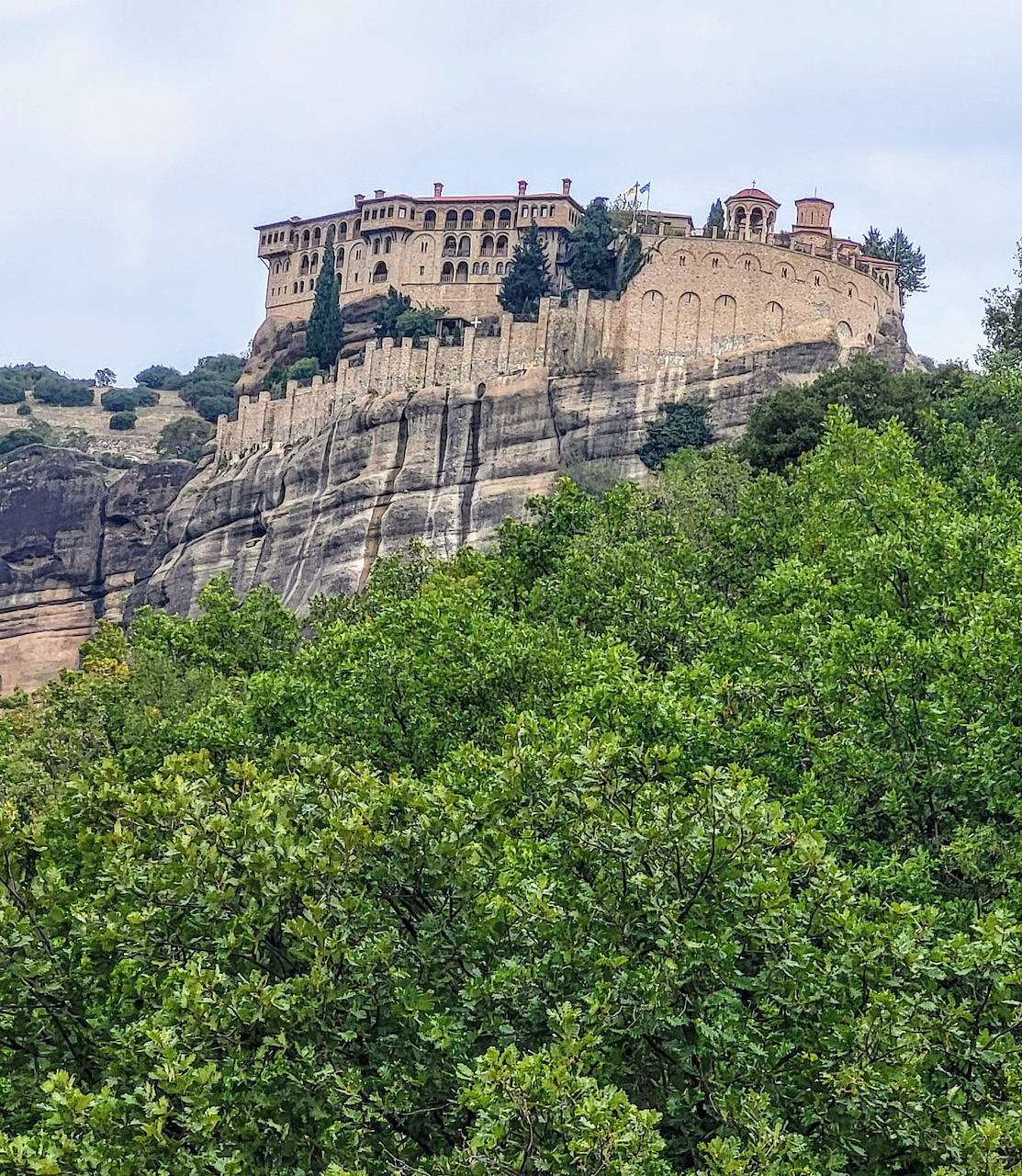 Stone monastery atop a sheer rock cliff in Meteora, Greece, with green trees in the foreground