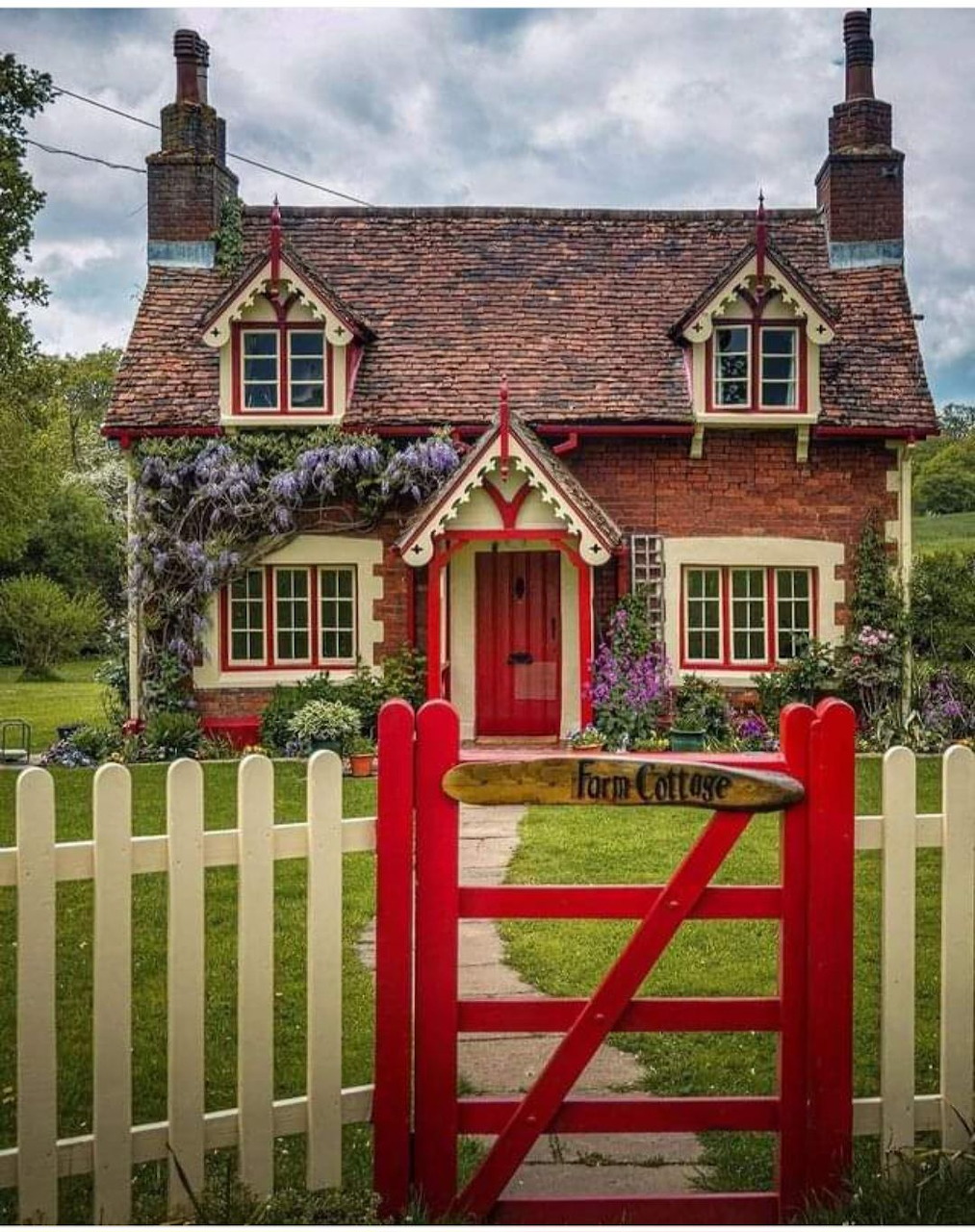 Red brick cottage with white fence, red gate and door, purple flowers climbing walls, green garden