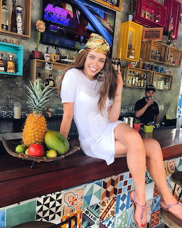 Smiling woman in white dress sitting on a colorful tiled bar counter with tropical fruits beside her
