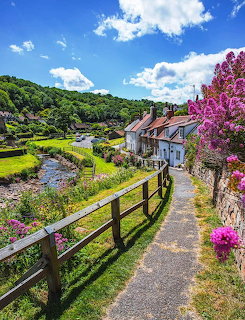 Stone cottages beside a stream with wooden fence, green valley, and purple blossoms under blue sky