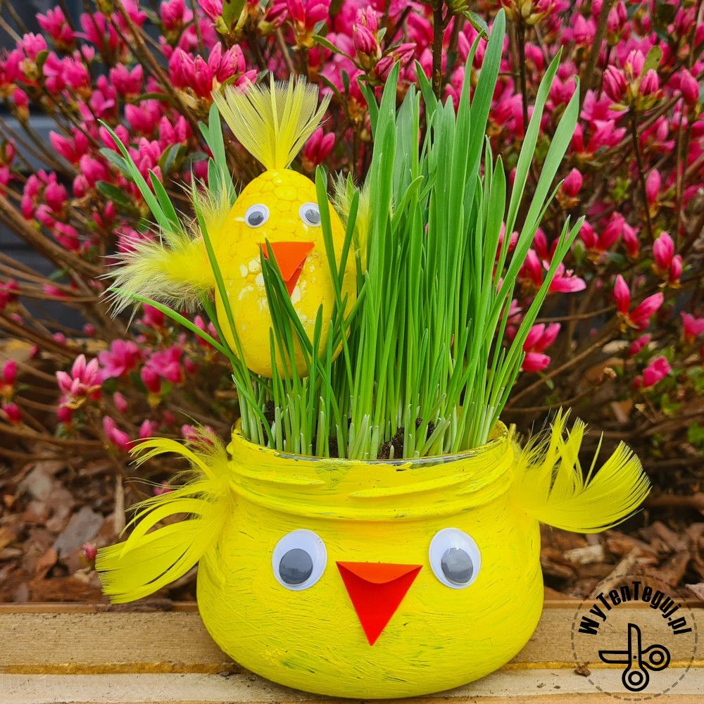 Yellow chick-shaped planter with googly eyes holding green grass and a toy chick against pink flowers