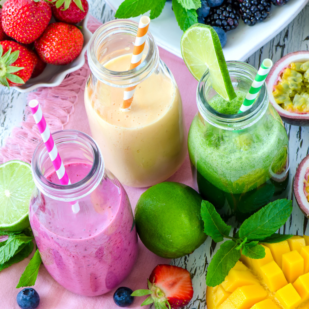 Three colorful smoothies with fresh berries, mango, lime and tropical fruits arranged on white wood background