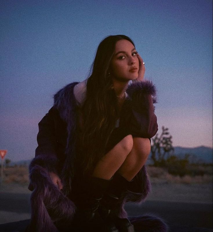 Young woman in purple fur coat sitting outdoors at dusk with desert landscape and twilight sky behind her