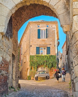 Stone archway framing a coblestone alley with sandstone buildings and blue shutters in a Mediterranean village
